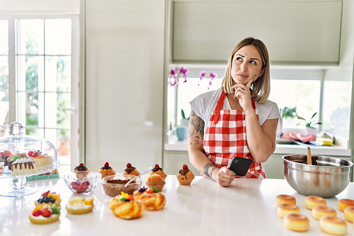 Young blonde woman using smartphone wearing apron cooking homemade pastries serious face thinking about question with hand on chin, thoughtful about confusing idea